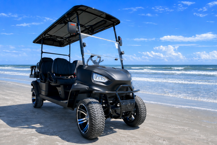 Black golf cart on a sandy beach with blue sky and ocean waves.