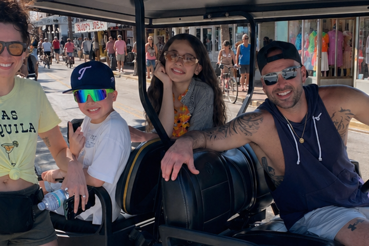 Smiling family on a golf cart in a sunny street, wearing casual clothes and sunglasses.