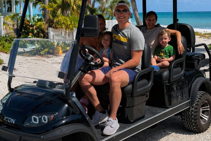 Family of five smiling on a golf cart by the beach.