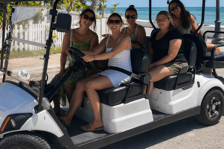 Five people in a white golf cart by the ocean, smiling on a sunny day.