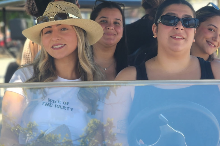 Four women smiling and seated in a golf cart, with one wearing a straw hat.