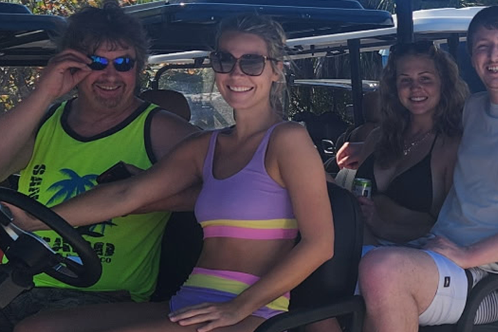 Four people sitting on a golf cart, smiling and wearing casual summer outfits.