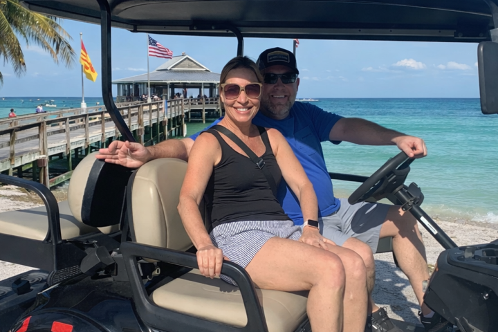 Two people smiling on a golf cart near a beach with flags and a pier.