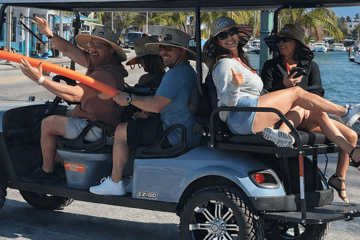 Group of people riding a golf cart near a waterfront with a rooster nearby.