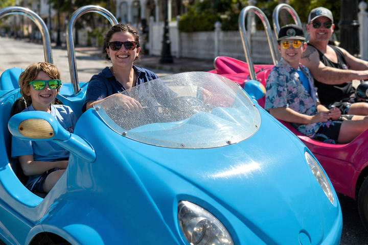 Group of people in blue and pink open-top cars on a sunny street.
