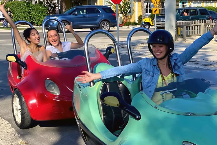 Three smiling people driving colorful, open-top mini cars on a sunny street.