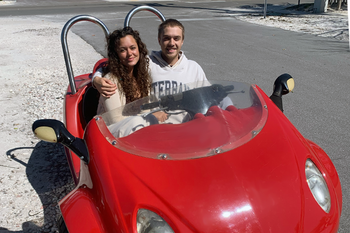 Two people sitting in a red dune buggy on a sunny day.