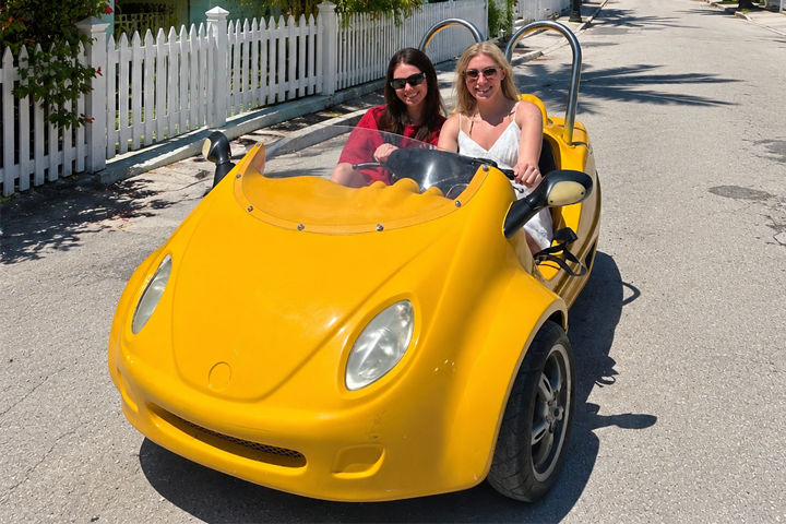 Two women smiling in a yellow open-top car on a sunny street.
