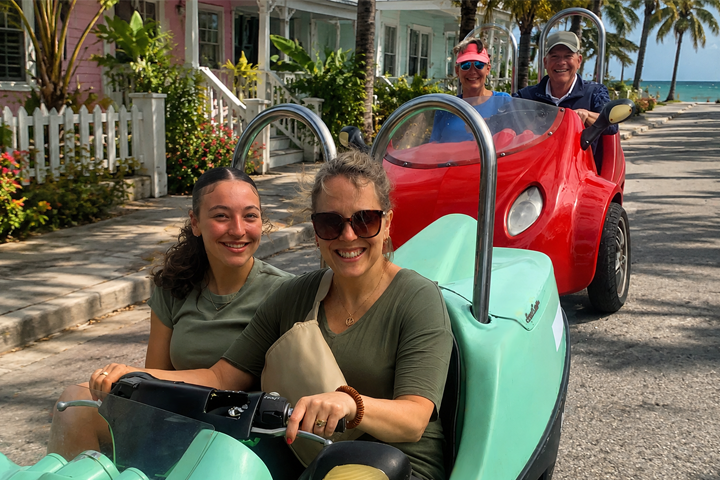 Two women in a small car lead two people in a red car on a palm-lined street.