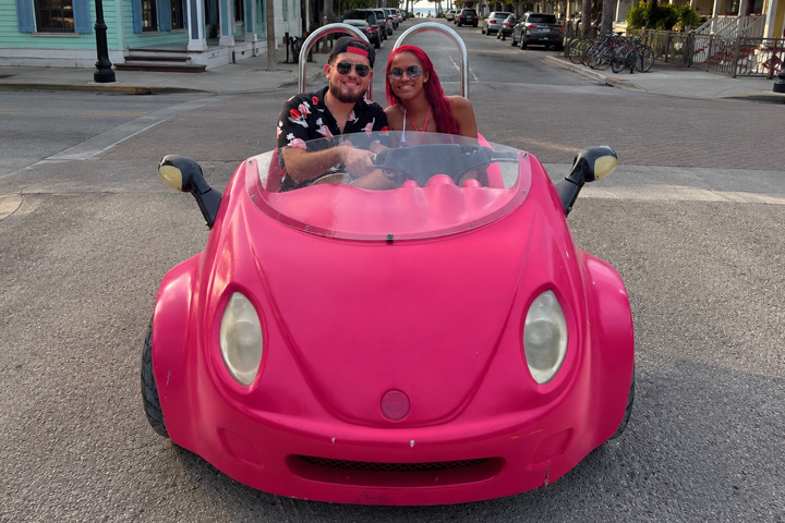 Two people in a small pink convertible car on a street.