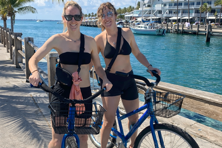 Two women in swimsuits with bicycles by a waterfront.