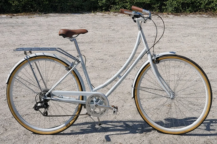 Classic white bicycle with brown seat and handles on gravel ground.