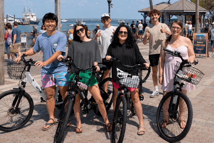 Group of smiling people with bikes near waterfront on a sunny day.
